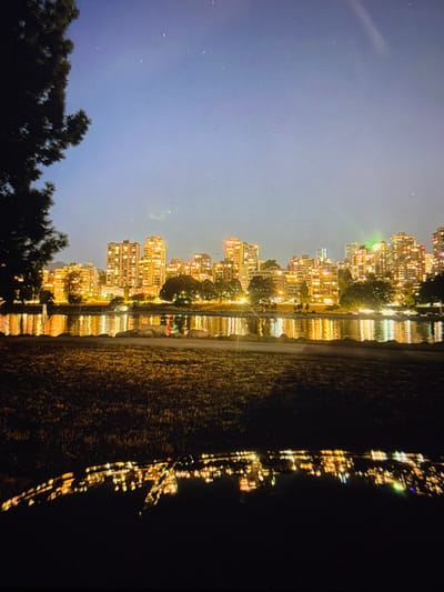 A nighttime urban landscape depicts the illuminated skyline of Vancouver, British Columbia, Canada, viewed across False Creek from a park-like setting. The location is within the Metro Vancouver Regional District, which includes Electoral Area A as an unincorporated area, though the prominent cityscape depicted is Vancouver proper.
The dark blue-grey sky above the city contains faint specks of light and a subtle, curved atmospheric streak. The immediate foreground features a dark, highly reflective surface, likely a vehicle hood, showing an inverted, distorted reflection of city lights. Above this, a strip of dark, unmanicured grass is partially illuminated by ambient light.
The midground consists of a calm body of water, likely False Creek, displaying numerous shimmering reflections of city lights in predominantly golden-yellow, with accents of red and green. The background showcases a dense array of multi-story buildings with many illuminated windows, primarily in warm yellow-orange light. Dark tree silhouettes are interspersed among the buildings and along the distant shoreline. A distinct bright green light source is visible on a structure towards the right of the skyline. No individuals, discernible actions, or interactions are visible.