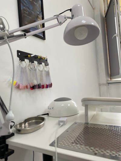 This is a close-up, eye-level, indoor photograph of a nail technician's workspace in Akyar, Russia. The dominant objects are a white desk, a white nail lamp, and a white UV lamp. Several nail polish sample fans are displayed on a black wall-mounted rack. A metal tray sits on the desk, and a metal grate with a built-in hand rest is also visible. The background shows a white wall and a window with a grey blind. The overall impression is one of a clean and organized professional setting.