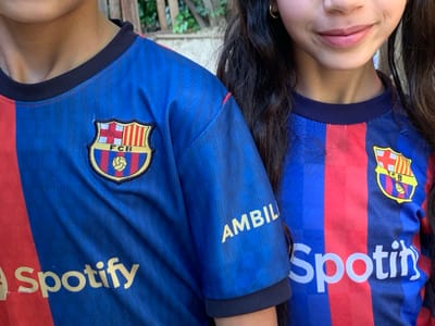 Two individuals, appearing young, are captured in a close-up outdoor shot, identified as being in Sidi Slimane Moul Al Kifane, Morocco. Both are wearing matching blue and red vertically striped FC Barcelona football jerseys with dark blue ribbed collars.
The individual on the left, visible from the base of the neck down to the lower torso and left upper arm, has a light skin tone. Their jersey features an embroidered FC Barcelona crest on the left chest. Below the crest, the "Spotify" logo is screen-printed in gold/yellow text on the lower left torso. On the right sleeve, the word "AMBIL" is visible in yellow text.
The individual on the right, visible from the mid-nose down to the lower torso and right upper arm, has long, dark, wavy hair. A small gold hoop earring is visible in their right ear. Their lips are closed in a slight, faint smile. Their jersey also displays an embroidered FC Barcelona crest on the left chest. The "Spotify" logo is screen-printed in white text on the lower right torso, and the word "AMBIL" is partially visible on the left sleeve.
The background behind the left individual features a light brown, possibly wooden, textured surface. Behind the right individual, dense green foliage is visible, suggesting an outdoor, possibly garden or patio, setting. The scene is evenly lit by natural light.