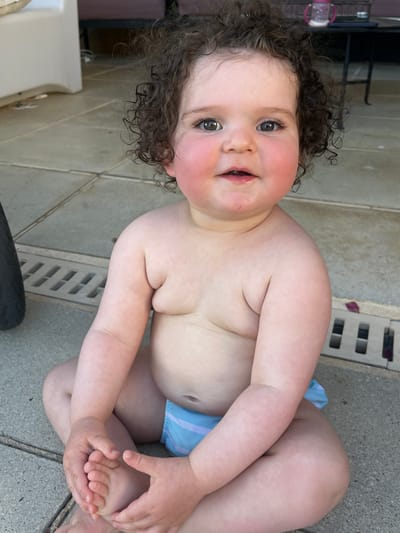 A young child with curly brown hair and rosy cheeks sits cross-legged on a tiled patio. The child is wearing a blue striped diaper. To the left of the child, a black tire is partially visible. In the background, there is a light-colored structure, a dark table with a pink sippy cup, and a drain grate. The scene appears to be outdoors, with sunlight illuminating the child. The location is identified as Saint-Galmier, France.