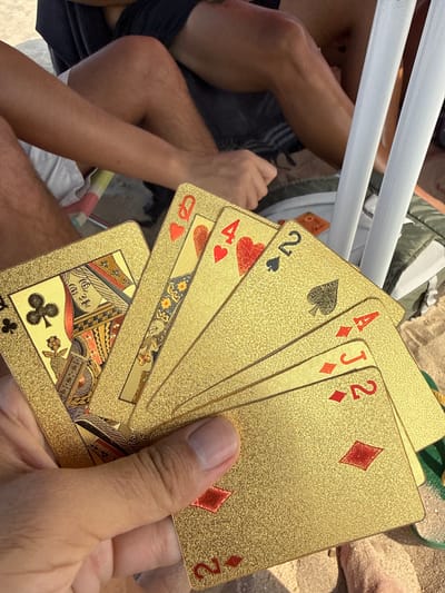 The image shows a person holding a fan of gold-colored playing cards on a sandy beach. The cards visible are the Queen of Hearts, the 4 of Hearts, the 2 of Spades, the Ace of Diamonds, the Jack of Diamonds, and the 2 of Diamonds. In the background, two people are seated on the sand, wearing dark shorts and one is wearing a white t-shirt. One person's legs are visible, showing tanned skin and hair. White plastic chairs are also visible in the background. The scene is consistent with a beach setting in Costa da Caparica, Portugal.