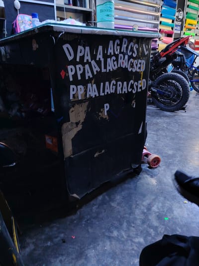 This image captures an indoor scene, likely a workshop, garage, or small business in Trujillo, Peru. The central focus is a dark-colored, makeshift counter, possibly constructed from cardboard or wood, showing signs of wear and tear.
On top of the counter, there's a light blue water bottle of the "BENEDICT" brand, clearly labeled "SIN GAS" and "3L" (3 liters) with "EXQUISITA AGUA" (exquisite water). Beside it is a child's lunchbox featuring characters from Disney's "Frozen," and some white, possibly fluffy, material. A small white object, possibly a hook or a light fixture, is attached to the wall above the counter.
The front of the counter is adorned with white, somewhat haphazardly applied letters and red diamond shapes, reading: "DPAA AGRCS", "PPAA LAG RACSED", and a repetition of "PPAA LAG RACSED", with a single "P" below. These appear to be decorative or informational, though their meaning is unclear.
In the background, to the right, parts of a black and red motorcycle are visible, with a blue object (possibly another bike or a fan) behind it. The motorcycle's rear wheel prominently displays a "Pornhub" sticker. Behind these, shelves are stacked with numerous rolls of colorful vinyl or similar material in various shades of blue, green, yellow, pink, purple, and silver, suggesting a business dealing with vehicle wraps or signage. A "Wi-Fi" sign and text mentioning "12 PERSONAS" (12 people) are also visible on the shelf unit.
The floor is a dark, possibly concrete, surface. A small pink child's scooter is partially visible near the base of the counter. A person's foot in a black shoe and another dark object are seen in the bottom right corner. The lighting suggests an indoor setting, possibly during the day or under artificial light.