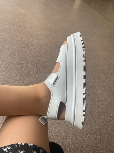 This media file features a close-up view of a white platform sandal on a person's leg. The sandal has a thick, white sole with a rugged tread pattern. The sandal's straps are also white and appear to be made of a smooth material, possibly faux leather, with a metallic detail on one of the straps. The person is wearing sheer, nude-colored stockings or tights. The background is a mottled brown surface, likely a floor or carpet, with visible texture. The lighting is even, suggesting an indoor setting. There are no discernible external location cues, time of day indicators, or weather information. The image focuses on the sandal and the leg, with no visible facial expressions or clear emotional context. No text is present in the image.