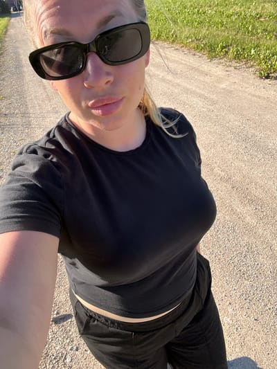 A woman is standing outdoors on a gravel path. She is wearing a black t-shirt and black pants. She has blonde hair and is wearing black sunglasses. The background shows green grass and trees. The location is identified as Zamosh'e (Zaklinskoe S/P), Russia.