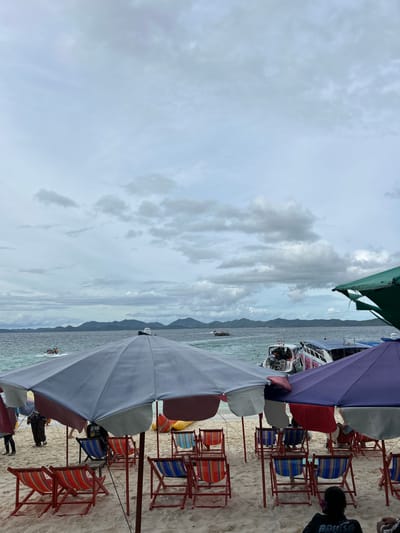 The image depicts a beach scene with several beach chairs and umbrellas set up on a sandy shore. In the background, the ocean is visible with several boats and a distant coastline with mountains under a cloudy sky. The scene appears to be in Tambon Phru Nai, Thailand, suggesting a tropical coastal location. There are a few people visible, some sitting in chairs and others walking. The overall atmosphere seems relaxed, though the cloudy sky might indicate a change in weather.