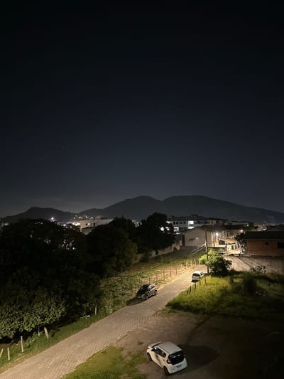 The image shows a nighttime view of a scene in Florianópolis, Brazil. The left side of the image is dominated by a dark expanse of water, likely the ocean or a bay, with a few faint stars visible. Adjacent to the water is a dark mountain range silhouetted against the night sky. On the right side of the image, a residential area with illuminated houses and streetlights is visible. A paved road runs alongside a grassy area with parked cars. The lighting is dim and uneven, with pools of light from streetlights and building illumination. The time is night. No people are visible.
