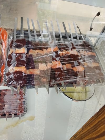 In Ala-Buka, Kyrgyzstan, skewers of raw meat, a mix of dark red and pale orange pieces, are displayed on metal skewers, covered in clear plastic wrap. The skewers are arranged on a metal tray placed inside a refrigerator or display case, with a white wire shelf visible in the background. A small portion of a glass of green liquid is also visible. The environment appears to be a food establishment.