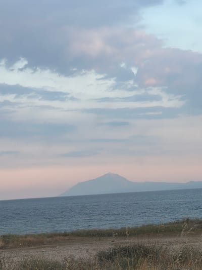 A tranquil scene unfolds across the image, depicting a vast expanse of deep blue water meeting a distant, hazy shoreline. A solitary, conical mountain rises majestically from the landmass on the horizon, its peak shrouded in soft, ethereal clouds that transition to a pale pink and lavender hue at the lower edges. The sky above is a canvas of muted blues and grays, punctuated by wispy clouds that create a dreamlike atmosphere. In the foreground, a strip of dry, grassy terrain borders the water, its muted tones suggesting a coastal or arid environment. The overall impression is one of serene, natural beauty, with the soft light and atmospheric haze contributing to a peaceful and contemplative mood. The location is identified as an unspecified area in Greece.