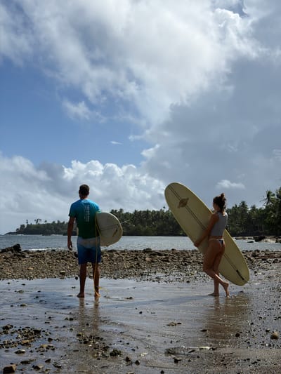 A man and a woman are walking on a wet, rocky beach, each carrying a surfboard. The woman is on the left side of the frame, carrying a light-colored surfboard on her head. The man is on the right side, carrying a surfboard on his shoulder with a yellow leash. The background includes a cloudy sky, the ocean, and a line of trees. The words "TAY HO 2024 HALF MARATHON" are on the man's shirt. The location is the beach in Santa Monica, Philippines.