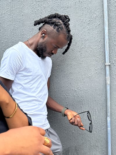 The image depicts a Black man with dreadlocks and a beard, wearing a white t-shirt, lying on what appears to be a gray concrete surface. He is holding a pair of black sunglasses in his right hand. A metal pipe runs horizontally across the top of the frame. In the foreground, partially obscuring the man, is another person's hand with a gold ring and a gold bracelet on their arm. This could be located in the city of Ojomu, Nigeria, but that cannot be determined by the image alone.