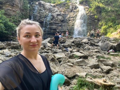In Peshcherka, Russia, a woman with blonde hair and green eyes takes a selfie in front of a rocky waterfall with a group of people in the background. The woman is wearing a black dress and a gold necklace. She is holding a blue inflatable object in her right hand. The waterfall is cascading down a rocky cliff face, surrounded by lush green foliage. Several people are visible near the base of the waterfall, some are posing for photos, while others are sitting or standing around. The ground is covered in rocks and pebbles, with some patches of grass and other vegetation. The overall scene depicts a scenic natural setting during a daylight excursion.