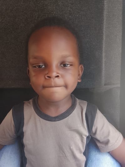A young child with dark skin and short hair is centered in the frame. They have a slightly furrowed brow and a neutral expression with pursed lips. The child is wearing a tan T-shirt with black sleeves. They appear to be sitting, with a glimpse of blue denim visible below. The background is a textured, dark gray surface. The lighting is slightly uneven, with shadows across the top of the child's head.