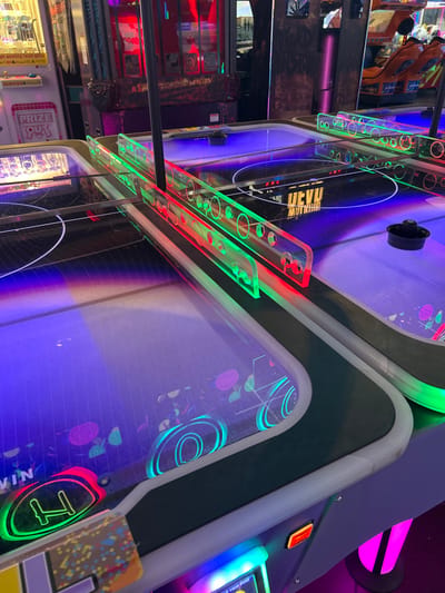 The image captures a vibrant, neon-lit arcade scene, likely within an entertainment center in Agios Dimitrios, Greece. The primary focus is on two side-by-side air hockey tables, viewed from an elevated, angled perspective. The tables glow with intense artificial lighting, casting purple and blue hues across their surfaces, creating a futuristic and energetic ambiance.

Transparent acrylic barriers, equipped with embedded red and green LED lights, separate the playing areas of the tables. These lights create dynamic streaks of color across the frame. One black air hockey puck rests on the surface of the top table, ready for play. The playing surfaces are adorned with swirling circular patterns and halftone-like designs in shades of cyan and magenta.

In the background, other arcade machines are partially visible, contributing to the bustling atmosphere of the venue. On a machine in the lower-left background, the text "PRIZE OUT" is visible. A reflection on the surface of the table further back displays the inverted text "LUCKY WINNER". On the front air hockey table, near what appears to be a score display, stylized green text reads "WIN," accompanied by a circular symbol resembling an 'H'.

The dim ambient light of the arcade, contrasted with the bright, colorful illumination from the machines, suggests an evening or nighttime setting. No people are visible in the frame, allowing the intricate details and glowing elements of the arcade equipment to take center stage. The scene evokes a sense of modern recreational fun.