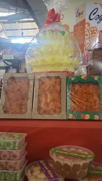 In Akyar, Uzbekistan, this photograph captures a market stall displaying a variety of confections. Dominating the upper portion of the image is a large, yellow, Christmas tree-shaped candy arrangement, intricately wrapped in clear cellophane and adorned with a red and gold bow. To the left, a sign in what appears to be Uzbek reads "Nozik did egalari uchun," suggesting it is for discerning tastes. Below this, three cardboard boxes, each filled with a translucent orange candy, are arranged side-by-side. The first two boxes are plain cardboard, while the third has a decorative green border with cutouts of fruit. In the foreground, several clear plastic containers are stacked. The top three containers are filled with pastel-colored, flower-shaped sweets, green, pink, and orange. Below these, a round container holds lighter colored cookies or candies, with a purple and green label that says "Qo'qon Halva" which is a type of halva made in Kokand. Another similar container is partially visible to the right. The market stall is set against a backdrop of overhead structures and what appears to be shelving units in the distance, suggesting an indoor or covered market environment.