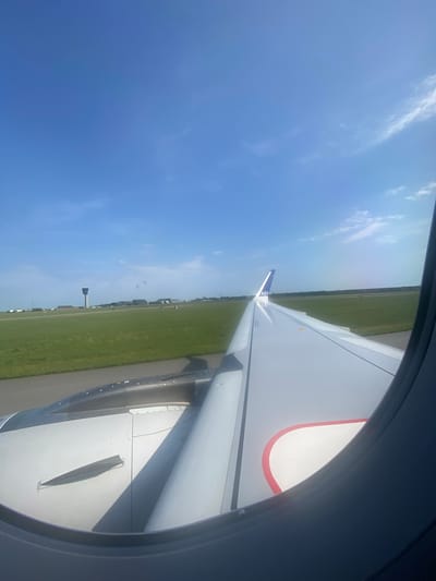 View from an airplane window. The wing of a SAS plane is visible with a blue and white tail. Below, a green grassy field can be seen with a runway and some buildings in the distance. The sky is blue with some clouds. The location appears to be near Copenhagen Airport, in Tårnby, Denmark, based on the surrounding landscape and the airline.