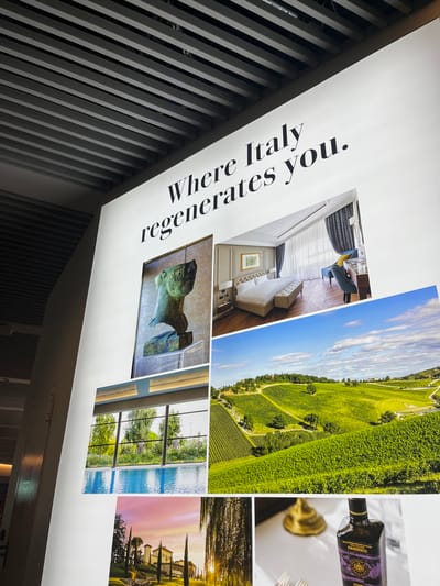 A large, brightly lit promotional wall display, featuring the prominent black serif text "Where Italy regenerates you.", is observed in a modern indoor environment located in Bologna, Italy. The display is mounted on a white wall, beneath a slatted grey ceiling. Six distinct photographic panels are arranged beneath the text. These panels include, from top-left to bottom-right: a bronze bust of a classical head, a luxurious hotel bedroom with a large white bed and elegant furnishings, expansive green vineyards under a partly cloudy blue sky, an indoor swimming pool with large windows overlooking trees, a Tuscan-style villa with cypress trees at sunset, and a close-up of a dark bottle of "OLIO EXTRAVERGINE DI OLIVA PANDESCO" on a white tablecloth alongside dining items. No individuals are present in the scene.