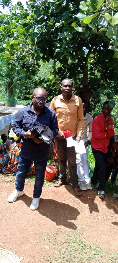 A group of four men stand on a dirt surface under a tree. The first man wears a blue shirt, jeans, and white shoes. He holds a cap and phone. The second man wears an orange shirt and brown pants, holding a megaphone. Behind them is a man in a white shirt and pants, and another in a red shirt and black pants with sandals. Some people sit and clothes hang in the background.