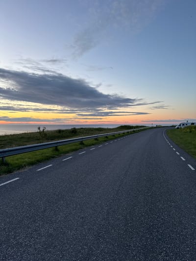 The image shows a coastal road at sunset in Paldiski, Estonia. The road is asphalt with white dashed lines. To the left of the road is a metal guardrail with green grass and some small trees. Beyond the grass is a body of water, likely the Baltic Sea, with a colorful sunset sky above it. To the right of the road are more grassy hills and two white campers parked. The sky transitions from orange and yellow near the horizon to blue and gray with scattered clouds higher up. There are no people visible in the image.