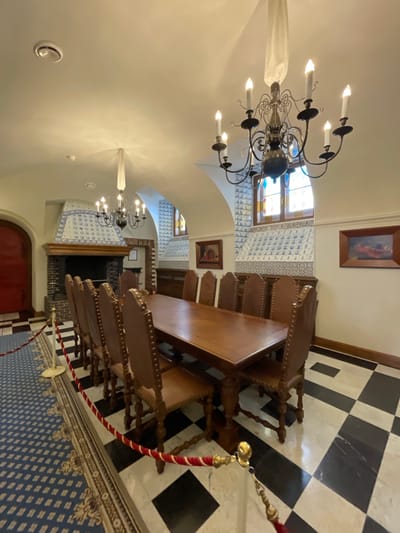 The image depicts a grand dining hall, likely within the Konstantinovsky Palace in Strelna, Russia. The room features a large, ornate wooden table surrounded by high-backed, upholstered chairs. Above the table, a vintage chandelier with multiple candle-shaped lights hangs from the ceiling. The walls are adorned with decorative blue and white tilework, and two framed paintings are visible. A large fireplace with a tiled surround dominates one wall. The floor is a striking black and white checkered pattern. Red and gold rope barriers are set up, suggesting the area is either on display or protected. The overall impression is one of historical elegance and preserved opulence.