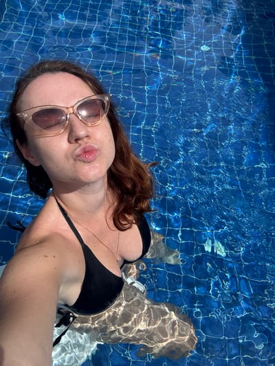 A woman in a black bikini and large cat-eye sunglasses is in a swimming pool, puckering her lips as if for a kiss. She has reddish-brown hair and is looking towards the camera. The pool water is a vibrant blue, with sunlight reflecting off the surface and creating a checkered pattern from the pool tiles. The woman is partially submerged, with water droplets visible on her skin and hair.