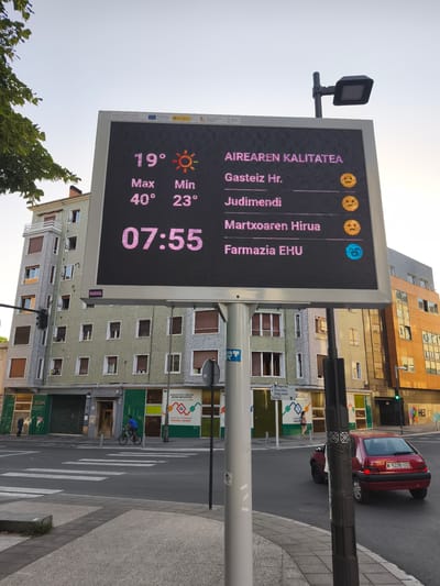 This media file is a photograph of a digital display board showing air quality information in Vitoria-Gasteiz, Spain. The display board is mounted on a pole at a street intersection, with buildings and trees in the background. The sky is clear and appears to be late afternoon or early evening.

The digital display shows the current temperature as 19°C, with a maximum temperature of 40°C and a minimum of 23°C. It also displays the time as 07:55, though this is likely an error given the lighting conditions. The air quality is indicated for different locations: "Gasteiz Hr." (Vitoria-Gasteiz Hr.), "Judimendi", "Martxoaren Hirua", and "Farmazia EHU". Each location has an accompanying smiley face icon, suggesting varying levels of air quality. "Gasteiz Hr." and "Judimendi" have yellow smiley faces, while "Martxoaren Hirua" also has a yellow smiley face. "Farmazia EHU" has a blue smiley face, possibly indicating better air quality. The display board also has government agency logos and text in Basque.

In the foreground, a red Citroën car with the license plate "M-5236-UD" is driving through the intersection. A person on a bicycle is also visible, riding on the street. The surrounding buildings include a multi-story residential building and a commercial building with storefronts. One storefront has the text "BIZAN SERVICIOS SOCIALES DE BAGE" and "Informezion eta orientazka Información y orientación" visible, indicating social services. Another building has "HEI" written on it. The scene is a typical urban streetscape with traffic and pedestrians. The overall impression is of a functional display board providing useful public information in an everyday urban setting.