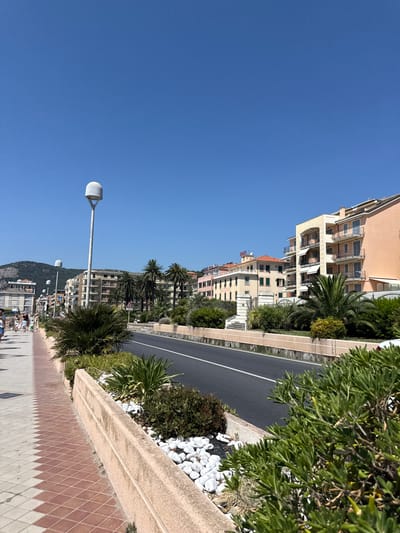 This image depicts a sunny day along a coastal road in Finale Ligure, Italy. On the left, a paved pedestrian walkway with a diamond pattern of terracotta and cream tiles lines the foreground. Beyond the walkway, a raised planter bed features lush green shrubs and white decorative stones. A street light with a modern, white globe design stands prominently.

Adjacent to the planter bed is a paved road with a white dividing line, indicating it's a two-lane road. Palm trees and other greenery are visible along the side of the road. In the background, several multi-story buildings with balconies and awnings are visible, suggesting a hotel or residential area. One building has a sign that reads "HOTEL". Further in the distance, a green, tree-covered hill forms the backdrop. A few people are walking along the pedestrian path, appearing to be enjoying the weather. The sky is a clear, vibrant blue, with no clouds present. The overall scene is bright and appears to be a typical summer day in a Mediterranean coastal town.