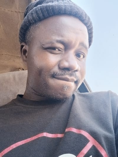 A close-up portrait, reportedly taken in Zonkwa, Nigeria, features an adult male with dark skin, a short dark mustache, and sparse stubble on his chin and jawline. He wears a dark grey ribbed knit beanie and a dark-colored, crew-neck t-shirt with a red diagonal stripe visible on its chest. The man's left eye is partially closed, his right eyebrow is slightly elevated, and his mouth is subtly pursed. The immediate background to his left is a light brown, textured fabric, consistent with a vehicle's interior panel. To his right, a clear, bright sky is visible through an opening, indicating an outdoor view from within a confined space.