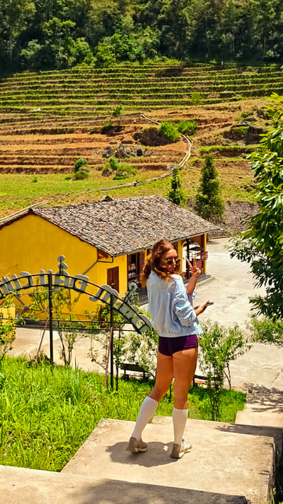 A woman is standing on concrete steps, facing away from the camera and slightly to the right, giving a peace sign with her left hand. She is wearing sunglasses, a light blue jacket, short purple pants, knee-high white socks, and tennis shoes. Behind her is a landscape featuring a yellow building with a brown-tiled roof. A white arched fence borders a grassy area in front of the building. Beyond the building, there are terraced fields ascending a hill, with dense green vegetation at the top. Additional greenery and a portion of a tree are visible on the right side of the frame. The setting is Lũng Cú Commune, Vietnam.