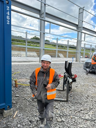 A man wearing a hard hat and an orange safety vest stands in front of a large blue shipping container at a construction site in Stróża, Poland. He holds a smartphone in his left hand and his right hand is in his pocket. The construction site features a steel structure with large beams and a gravel-covered ground. In the background, a van and some construction equipment are visible. The sky is partly cloudy.