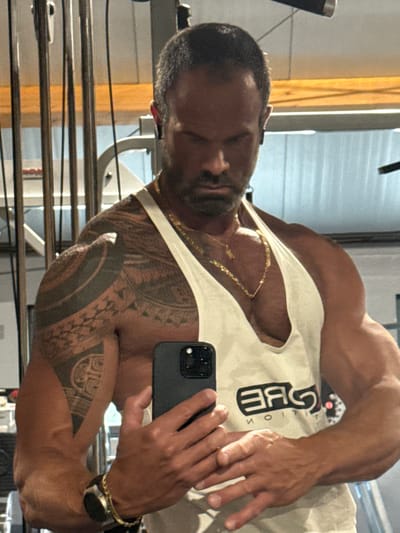 The image shows a muscular man with tattoos on his left arm and shoulder, wearing a white tank top, gold necklaces, and a smartwatch on his left wrist. He is holding a black smartphone in his right hand and appears to be taking a selfie in what looks like a gym. The background includes gym equipment such as weight racks and cables. The man has a beard and is looking down. The lighting suggests an indoor setting, likely at night or in a dimly lit area. There is no indication of the specific location within the city of Jemelle, Belgium.