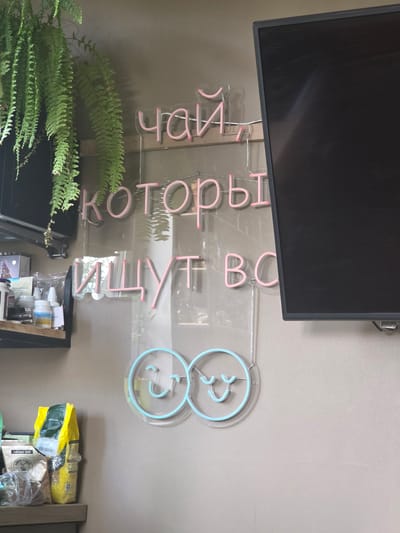 The image shows a wall with a neon sign. The neon sign spells out "ЧАЙ, КОТОРЫЙ ИЩУТ ВСЕ", which translates to "Tea that everyone is looking for." There are also two smiley faces made of blue neon lights. To the left of the sign, there is a shelf with various items, including bags of tea or coffee, and some bottles. A large fern plant hangs from above, partially obscuring the wall. A black television screen is visible on the right side of the image. The overall environment appears to be indoors, possibly a cafe or a shop. The location is identified as Brilliant, Russia.