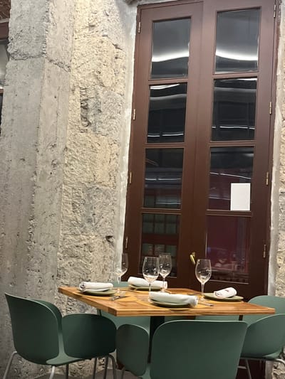 The scene shows an outdoor dining area located on the patio of a restaurant in Las Palmas de Gran Canaria, Spain, with a view of the nearby street. The patio is enclosed on three sides by a concrete wall with a large brown wooden door on one side and a large concrete pillar with chipped paint on the other. There are five green chairs around a table, two of which are occupied, and one of which is empty. The table has a wooden top and a metal base. On the table are four place settings with white napkins, silverware, and glasses. The table has a glass with a red liquid on it. There are four wine glasses, one of which is on the empty setting, and three of which are filled. The table is decorated with a wooden tray with a pattern of small, alternating light and dark squares. Two of the glasses on the table have a red liquid in them. The door appears to be made of two panels and has a brass handle and several hinges. The door is closed and the glass panels are slightly frosted. There are several other objects visible in the reflection in the glass door panels, but their details are not clear. The scene is lit by the sun shining through the glass panels of the door. The air temperature appears to be moderate. The image was taken on a clear day with minimal cloud cover.