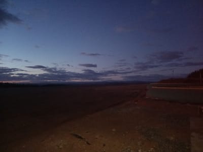 The image depicts an expansive, uncultivated landscape during late twilight in an unidentified area within the municipality of Santa Juana, Chile. The immediate foreground comprises dark, uneven ground, possibly dirt or gravel. Extending into the midground is a broad, flat, dark field or plain. Along the right side of the frame, a series of light-colored, tiered horizontal structures, resembling large pipes or retaining walls, are visible at the base of a densely forested incline. A single, slender dark pole ascends vertically behind these structures. In the far background, distant hills or mountains are silhouetted against a sky transitioning from deep indigo overhead to a lighter blue-gray near the horizon, accented by scattered clouds and a faint orange glow. No individuals, animals, vehicles, or active elements are present in the scene.