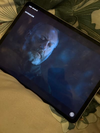 A tablet displays a close-up of a solemn, older man with a beard. The screen shows what appears to be a scene from a movie or television show, set in a dimly lit, perhaps underwater, environment. A time code and website, "egydead.com," are visible in the corner. An outward-pointing arrow icon is displayed in the upper right corner of the screen. The tablet sits on a patterned, green and white, bedsheet-like material.