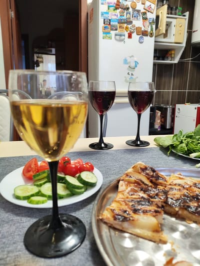 This image captures an indoor dining scene in Malaya Purga, Russia. The foreground features a clear wine glass with a black stem, filled with a golden-yellow liquid, positioned centrally and slightly out of focus. To its left, a white plate holds sliced red tomatoes and green cucumbers. To the right, a metallic platter presents several slices of a baked item, possibly a tart, with a caramelized or charred topping over a light-colored filling.

In the mid-ground, behind the prominent white wine glass, two clear wine glasses with black stems are filled with a dark red liquid, likely red wine. Further to the right, a white plate contains a variety of fresh green leafy vegetables, including spinach and possibly dill and green onions. The table surface is covered with a textured grey placemat or tablecloth.

The background reveals an open wooden door frame on the left, through which an indistinct reflection of a person is visible. On the right, a white refrigerator is covered with numerous colorful souvenir magnets and a Santa Claus decoration. Adjacent to the refrigerator, a white open shelf unit holds a microwave oven with its door slightly ajar. Above and below the microwave, small spice jars and bottles are visible. The wall behind features dark brown tiled surfaces. Parts of other kitchen appliances, including a dark metallic machine and a white and red appliance, are visible on the far right. The overall scene depicts a prepared meal set on a table in a domestic environment.