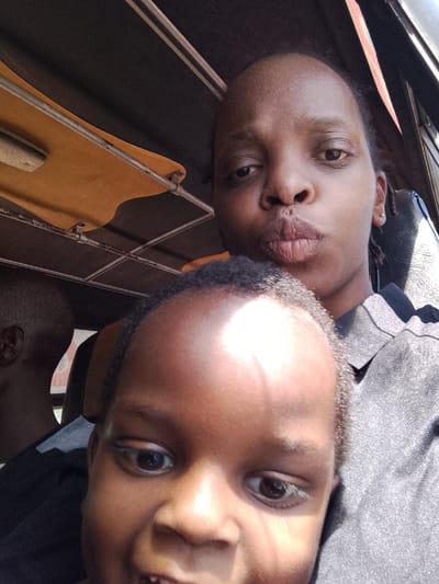 A woman and child are in a vehicle, possibly a bus or van. The woman is behind the child, who is close to the camera. The woman has dark skin, short hair, and is wearing a gray top. She is pouting her lips toward the camera. The child has darker skin and is smiling, revealing teeth. The vehicle interior has a yellow-tanned fabric lining with metal supports. Part of another person's head is visible to the left.