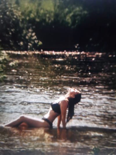 The image depicts a woman in a black bikini lying on her back in shallow water. Her head is tilted back, and her eyes are closed, suggesting a relaxed or serene emotion. Her dark hair is fanned out in the water behind her. The water is murky and appears to have a rocky or pebbly bottom. In the background, there is lush green foliage, indicating an outdoor, natural setting, likely a river or stream. The lighting suggests it is daytime, possibly a sunny day due to the bright reflections on the water's surface. The overall scene conveys a sense of peace and enjoyment of nature. There is no visible text in the image.