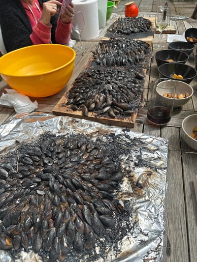 The image captures an outdoor food preparation scene in La Couarde-sur-Mer, France. Several wooden planks form a table surface on which the main subject of the image rests. Three wooden planks form a tabletop. Two sets of cooked mussels dominate the view. The first set is arranged on a wooden board, the second on a piece of aluminum foil. Next to the mussels, six ceramic bowls with fried potatoes are visible, along with three empty wine glasses. In the lower-left corner, a yellow plastic bowl and a partially visible person in a red sweater and black pants suggest active preparation. The background consists of a light-colored wooden wall.