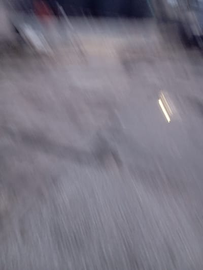 The image is a blurred, low-light photograph with a diagonal motion blur. It appears to be an outdoor scene, possibly a street or an open area. There are faint suggestions of structures or vehicles in the upper portion of the image, with some indistinct colors that could be red or dark blue. A bright, elongated light source is visible in the mid-right section, appearing as streaks due to the motion blur. The ground or surface is a muted, greyish tone with a texture suggestive of gravel or rough pavement. The overall impression is one of movement or a hasty capture in dim lighting conditions. Without more distinct features, pinpointing a specific location in Port Harcourt, Nigeria is not possible.