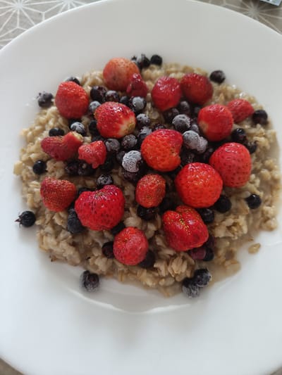 A white bowl of oatmeal is topped with fresh strawberries and blueberries. The oatmeal appears to be cooked with water or milk, giving it a creamy texture. The strawberries are bright red and plump, with some showing their green tops. The blueberries are dark blue and round, with a slightly frosted appearance, suggesting they may be frozen. A few of the berries have a light dusting of frost. The dish is presented on a white plate, which is placed on a patterned placemat with a geometric design in shades of grey and white. The lighting suggests it is daytime, possibly morning or afternoon, with natural light illuminating the food. The overall scene evokes a feeling of a healthy and delicious breakfast. The context of Vichuga, Russia is noted, but no specific visual cues from the image directly indicate this location. There is no visible text in the image.