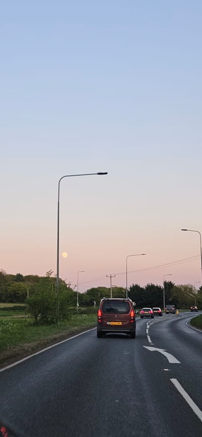 This image, when viewed in correct orientation, captures a road scene at either dawn or dusk in South Norfolk, United Kingdom. The sky transitions from a soft light blue at the top to a warm, peachy-pink near the horizon, where a pale, full moon is distinctly visible.
In the foreground, an orange-brown van-like vehicle, bearing the UK license plate "KY21 CRN," is positioned on the wet asphalt road with its red taillights illuminated. Ahead, several other vehicles are visible, also displaying their illuminated taillights, indicating traffic flow. The road surface appears damp, suggesting recent rain. White road markings, including a clear white arrow directing traffic to turn right, are visible on the asphalt.
Several tall, dark streetlights with curved arms extend upwards, silhouetted against the gradient sky. To the left of the road, green trees and bushes line the route, with open fields visible further into the distance. Utility poles and overhead lines are also present in the mid-ground. The scene conveys a tranquil moment on a road, likely during a commute as light fades or emerges.