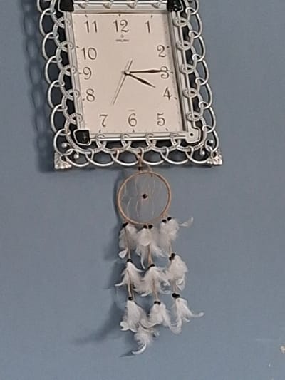 A light blue wall in the city of Selyanin, Bulgaria, features a wall clock mounted above a decorative hanging. The rectangular wall clock, positioned centrally in the upper frame, exhibits an ornate silver-toned metallic frame with a chain-link pattern and black embellishments at its corners. Its white dial displays black Arabic numerals (1-12) and black hour and minute hands, indicating approximately 2:15. The word "BULGARI" is inscribed below the '12' numeral. Directly beneath the clock, a decorative item hangs, comprising a light-brown circular hoop from which multiple strands descend. Each strand terminates in a cluster of small, white feathers, often secured by a dark bead at its origin. Shadows are visible on the wall, cast by the mounted objects. No people or interactions are present in the scene.