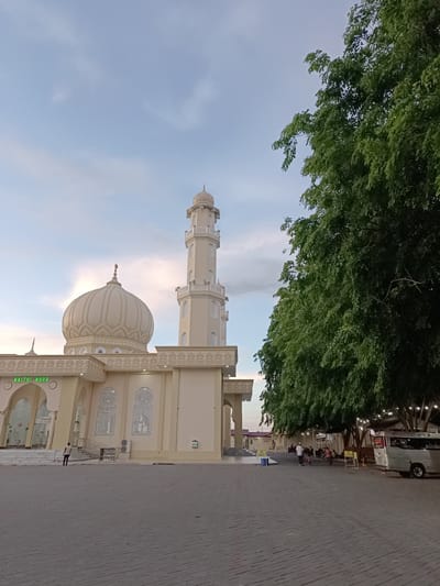 This image depicts the Baitul Huda mosque in Mee Tanjong Usi, Indonesia, during what appears to be the late afternoon or early evening. The sky is a soft blue with scattered clouds, suggesting fair weather.
The mosque is the central subject, characterized by its light-colored facade, a prominent dome, and a tall minaret. Intricate decorative patterns adorn the building's facade, particularly around the windows and along the upper ledges. The name "BAITUL HUDA" is clearly visible in green lettering above the entrance.
In the foreground, a paved courtyard stretches out, with gray interlocking pavers forming a textured surface. To the right, a large, mature tree with dense green foliage dominates the frame, its branches extending over a portion of the courtyard. Beneath the tree, an area is set up with lights, suggesting it may be a gathering or resting spot. Several people are visible in this area, some standing and others walking. A silver minivan is parked in this vicinity.
Further to the left, near the mosque entrance, a few individuals are also present, with one person walking away from the camera. The overall atmosphere appears calm and serene, possibly indicating a period after prayer services or a quiet time of day. The lighting suggests the sun is beginning to set, casting a gentle glow on the scene.