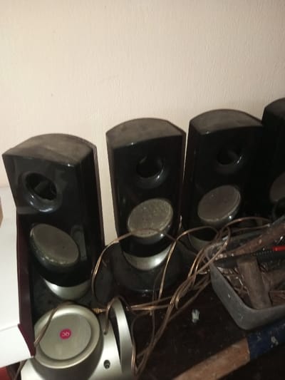 A group of black speakers sits on a surface, dusty with visible wear. The speakers have circular openings and smaller, textured centers. Wires snake around the base of the speakers. Next to the speakers is a plastic container filled with tools and what appear to be pieces of wood. A blue-painted object, possibly a hammer handle, is also visible. A white or silver base unit sits near the first speaker.