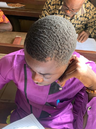 The image depicts an indoor scene in Abobo, Nigeria, with three individuals. The primary subject, a dark-skinned male, is viewed from a high angle, positioned in the foreground and center. He wears a purple long-sleeved shirt with black vertical accents and metallic circular buttons, and a blue pen is clipped to his left chest pocket. His short, dark hair exhibits a wavy texture, and his gaze is directed downwards towards a piece of white lined paper partially visible in the lower left. His left hand, adorned with a multi-beaded bracelet, rests against his jawline. Behind him, another dark-skinned male is partially visible, wearing glasses and a black shirt with gold patterned motifs. This individual is also looking downwards at a document on a wooden surface. To the far left, a third individual, only partially visible, wears an orange patterned garment. The environment consists of wooden desks or benches, suggesting an educational, meeting, or administrative setting.