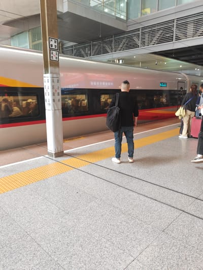 A high-angle shot captures a scene at a train station platform in Yuhuatai District, China, during the daytime. The main subject is a man standing with his back to the camera, wearing a black long-sleeved shirt, blue jeans, and white sneakers, with a black backpack slung over his shoulder. His hair is short and faded on the sides. He stands on the textured grey platform, which features a prominent yellow tactile paving strip near the edge.
To his right, other passengers are partially visible, including a woman in beige clothing with a red rolling suitcase, appearing to be waiting. Alongside the platform is a modern high-speed train, possibly a Fuxing Hao series, with a sleek silver-grey body, a red stripe along the bottom, and bright yellow accents. Reflections of people can be seen in its windows. A digital display on the train shows the number "5".
A tall, grey pillar stands between the man and the train, adorned with multiple warning signs. The signs, written vertically in Chinese, clearly state "严禁登" (Strictly forbidden to climb), "高压危险" (High voltage danger) accompanied by a lightning bolt symbol, and "禁止靠近" (No approaching/Keep off), the latter repeated twice. The station features a high, modern ceiling with visible ventilation structures and lighting. The overall atmosphere suggests a routine travel day, with people calmly waiting to board or depart.