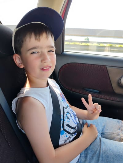 A young boy wearing a blue baseball cap and a white sleeveless shirt with a blue graphic print is seated in the back of a car. He is wearing denim shorts and a seatbelt. The boy is making a peace sign with his right hand and smiling at the camera. Sunlight streams in from the right, casting a shadow on his left shoulder. The car's interior is visible, including the dark upholstery of the seat and door panel with a red mesh pattern. Outside the window, a blurred landscape of trees and a highway barrier can be seen, suggesting the car is in motion on a sunny day. The image was taken in Orlovka, Russia.