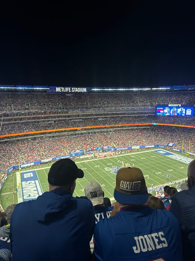 The image displays an overhead view from spectator seating within MetLife Stadium in East Rutherford, United States, during a nighttime American football event. In the immediate foreground, the backs of three individuals are visible. The person on the left wears a dark blue hooded top and a black baseball cap. The central individual wears a light-colored, patterned baseball cap with "NY" lettering. The person on the right wears a blue jersey with "JONES" printed in white on the back and a brown baseball cap displaying "GIANTS."

The football field below is illuminated and features white line markings, yardage numbers, and blue end zones, one of which explicitly shows "GIANTS" in white lettering. Multiple tiers of stadium seating, filled with numerous spectators, encircle the playing field. Above the uppermost seating deck, an illuminated sign clearly identifies the venue as "METLIFE STADIUM." A large video scoreboard on the right side of the stadium displays a "MOODY'S" logo and additional graphics. The sky above the stadium is dark, indicating a nighttime setting, with stadium lights providing primary illumination.