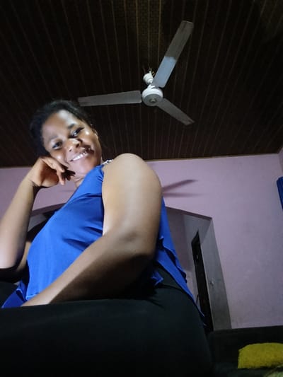 A low-angle, medium shot captures a Black woman in a blue top and black leggings, seated indoors. She rests her chin on her hand and smiles at the camera. Behind her, a ceiling fan is visible against a dark, wood-paneled ceiling. The walls are painted a light purple, and a doorway is visible on the right side of the frame. The scene appears to be in Yenegoa, Nigeria.