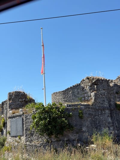 In Vranjina, Montenegro, on a clear, sunny day, the remnants of an ancient stone structure stand prominently against a bright blue sky. A tall, white flagpole with a red flag is positioned slightly to the right of the center of the frame, its fabric gently waving in the breeze. The stone ruins are weathered and overgrown with patches of green vegetation, suggesting a site that has endured for a considerable time. On the left side of the ruins, a weathered gray plaque is affixed to the stone wall, inscribed with text in what appears to be a foreign language. The foreground is characterized by dry, tall grass, with more verdant greenery and bushes growing around and on top of the stone walls. Power lines are visible stretching across the top of the image, indicating the presence of modern infrastructure near the historical site. There are no people visible in the image.